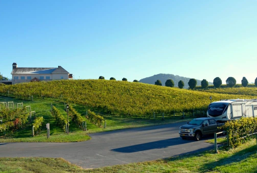A truck towing a fifth wheel sits in front of a large property overlooking a vineyard.