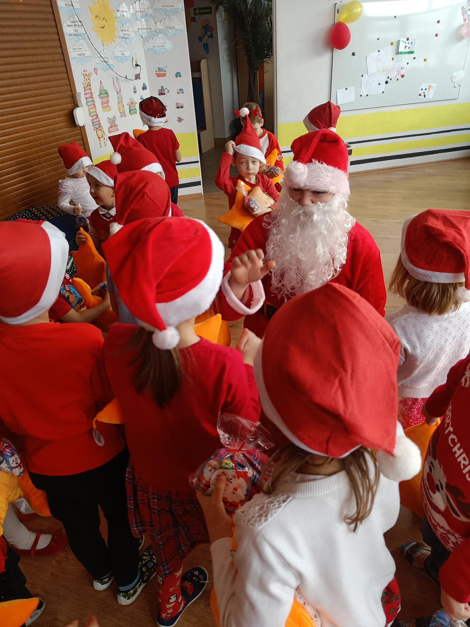 Mikołaj w przedszkolu | Wychowanek MOW przebrany za Mikołaja otoczony grupką dzieci w mikołajkowych czapkach.jpg