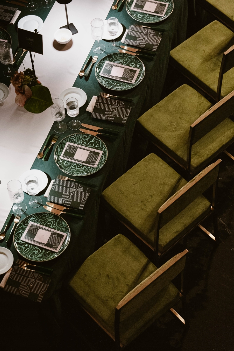 an overhead view of a long rectangular dinner table at a gala at Vibiana in Downtown Los Angeles. the table settings are white and emerald green, and the chairs are square with velvet moss green cushions. the chairs are angled.