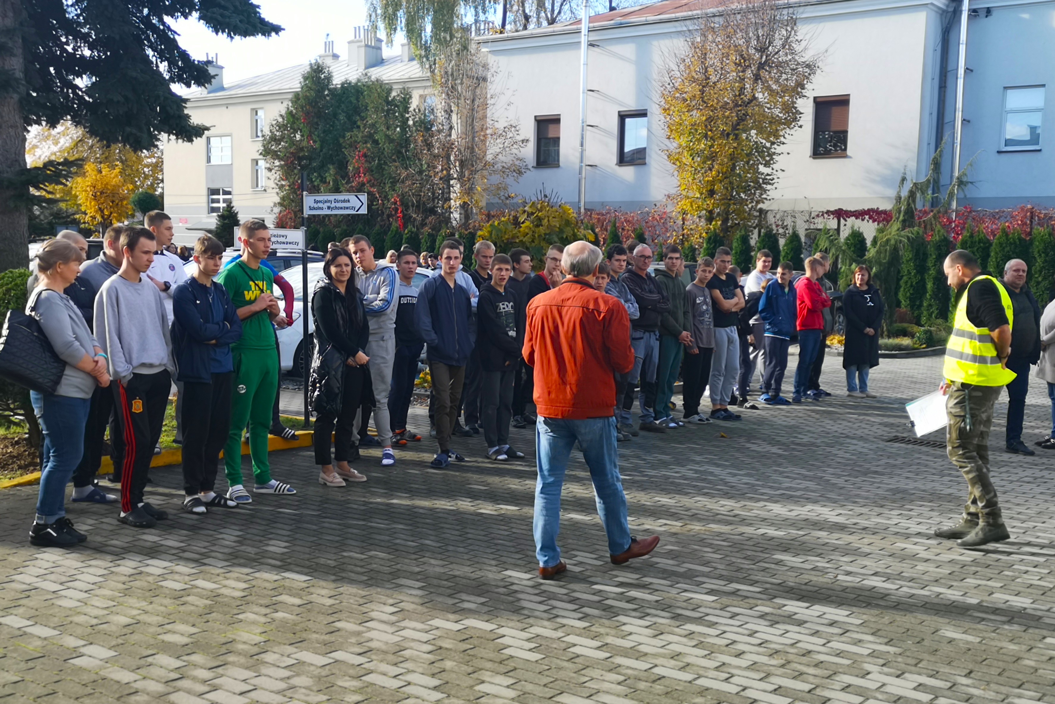 Próbna ewakuacja przeciwpożarowa | Wychowankowie MOW stoją klasami w dwuszeregu na wybrukowanym szarą kostką placu. Obok każdej klasy stoi nauczyciel. Przed chłopcami odwrócony tyłem instruktor BHP  oraz nauczyciel w kamizelce odblaskowej odpowiedzialny za ewakuację..jpg