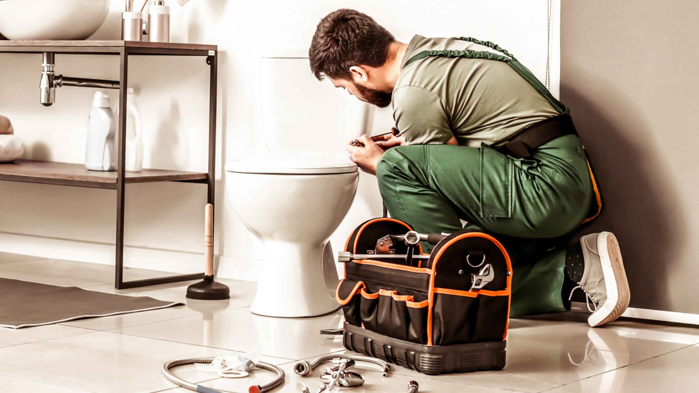 Plumber in green overalls, kneels beside a toilet. His tool bag, and plumbing parts are laid out beside him.
