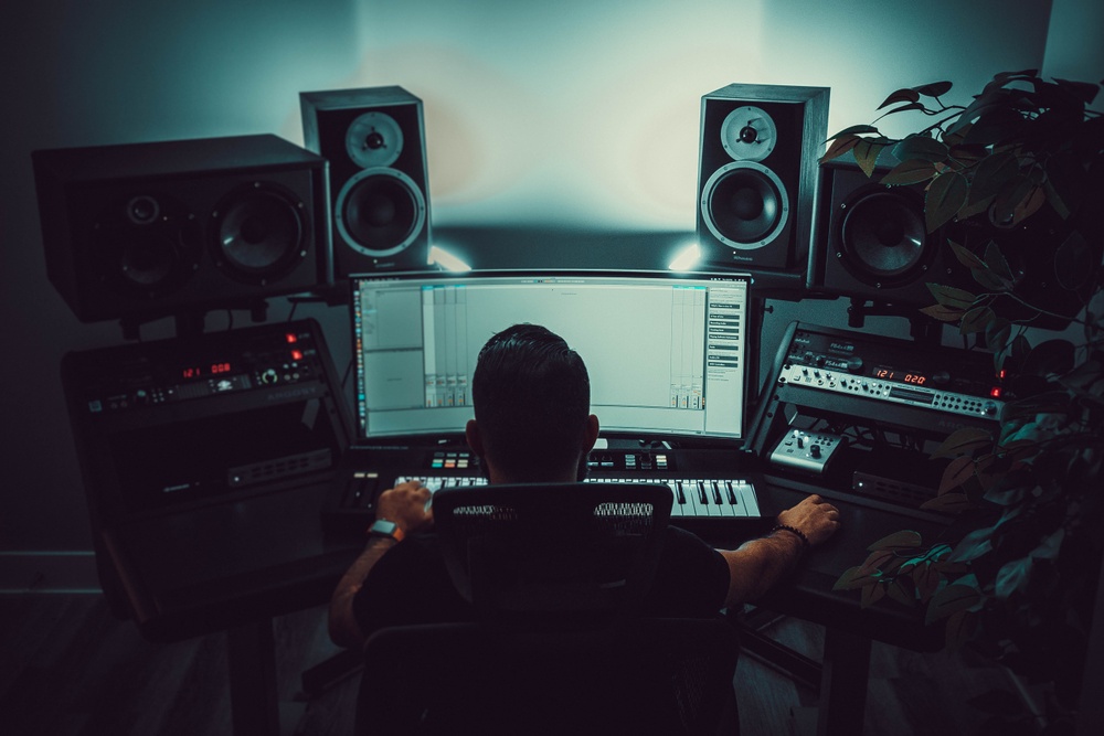 a person sitting in front of a computer producing music. There are computers, consoles and sound systems around the desk.