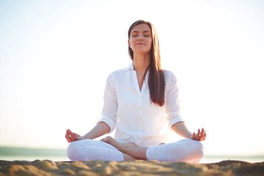 Young woman sitting cross legged by water in yoga position, eyes