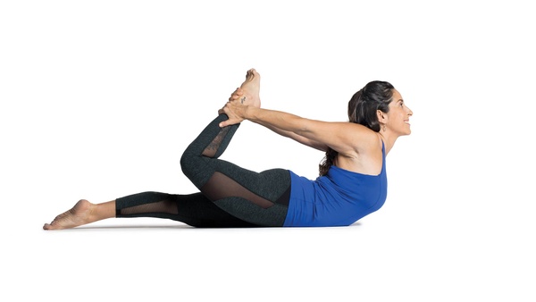 Flexible young female with curvy body exercising indoors, doing dhanurasana  or bow pose on yoga mat to strenghten back, open chest and shoulders,  improve digestion and stimulate reproductive organs Stock Photo