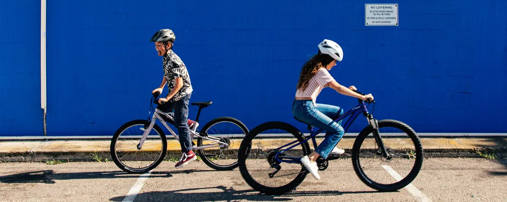 Two kids ride bikes past a blue wall