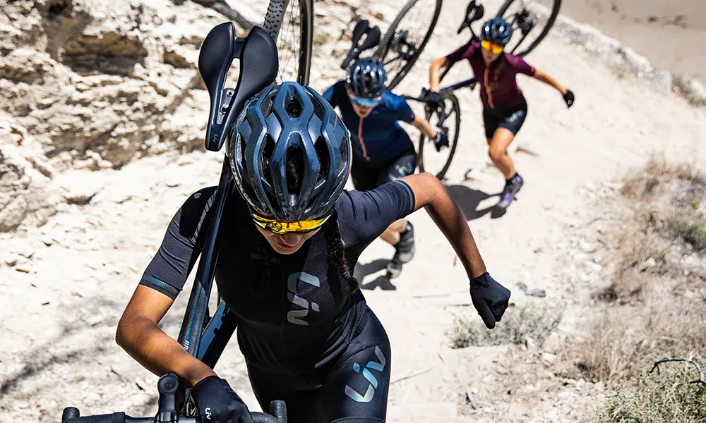 Three women carry cyclocross bikes up a sandy hill