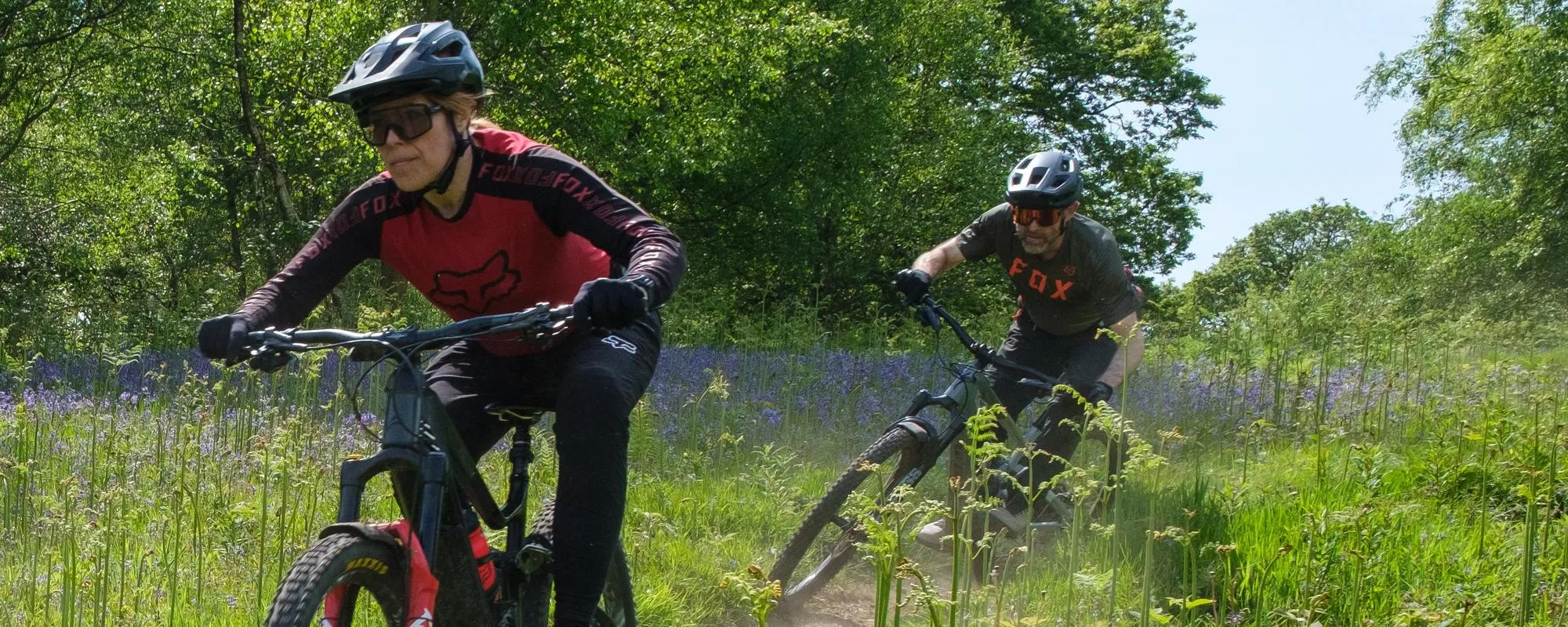 Two mountain bikers riding a track bordered with Bluebells wearing Smith Sunglasses and Fox clothing