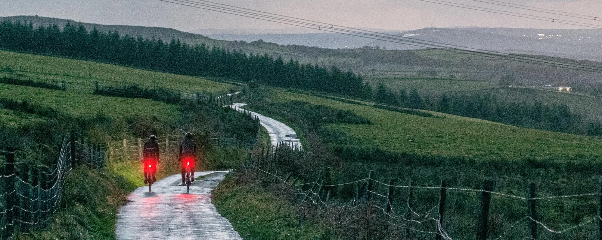 two road cyclists with red rear lights riding on country roads on a damp day at dusk