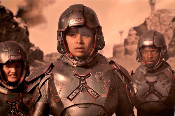 Two women and a man wearing futuristic grey space suits, facing the camera looking serious