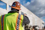 A construction worker wearing a hi-vis vest and a hardhat pointing to a wall being erected in the distance