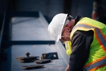 Two contractors wearing hi-vis vests and hard-hats looking down at a workbench