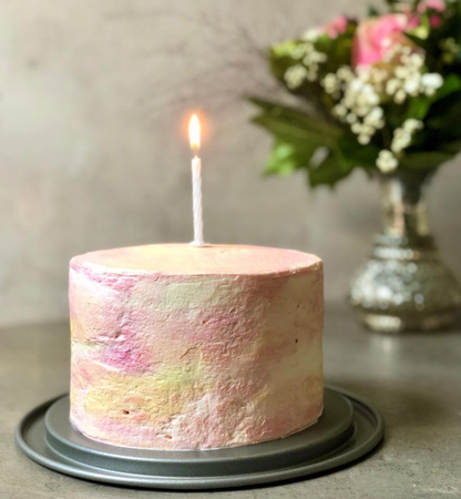 A cream covered birthday cake stained with natural plant-based food colorings and a single lit candle on top