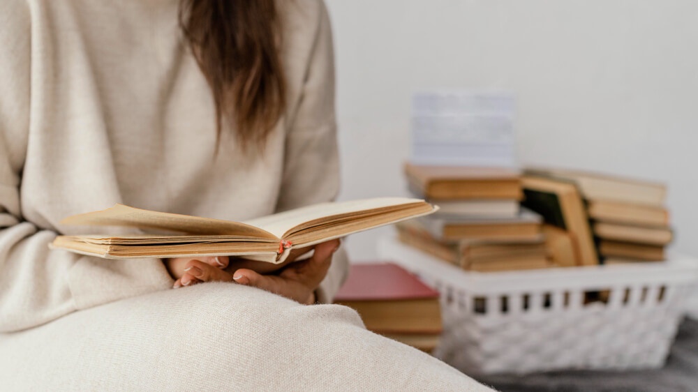 Seorang gadis tengah duduk membaca buku berlatarkan selonggok buku dalam bakul. 