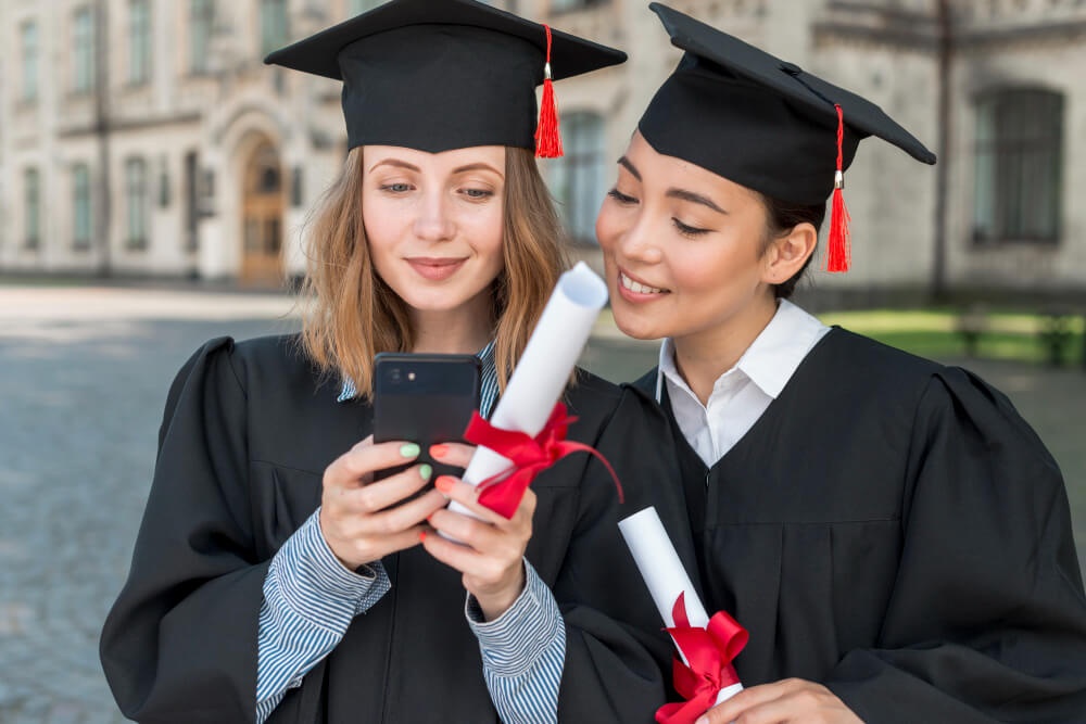 Dua orang graduan dengan pakaian dan gulungan ijazah membaca hantaran pada telefon bimbit. 