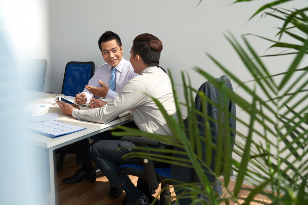 Two cheerful young businessmen are seated at a desk, engaged in a discussion and planning a work meeting