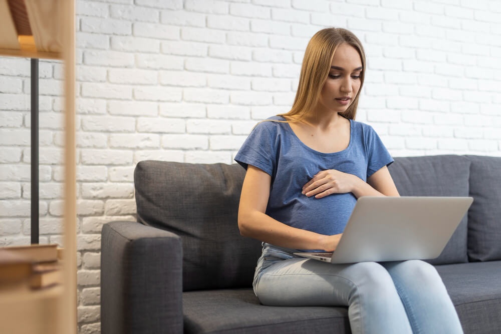 Wanita hamil duduk atas sofa sambil memberikan tumpuan kepada komputer riba