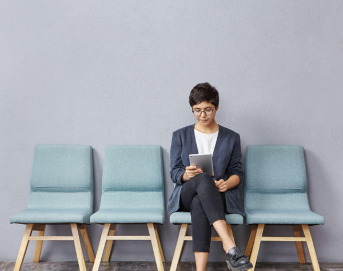 A young woman sits in a waiting area, reviewing a tablet and preparing to address selection criteria before her job interview