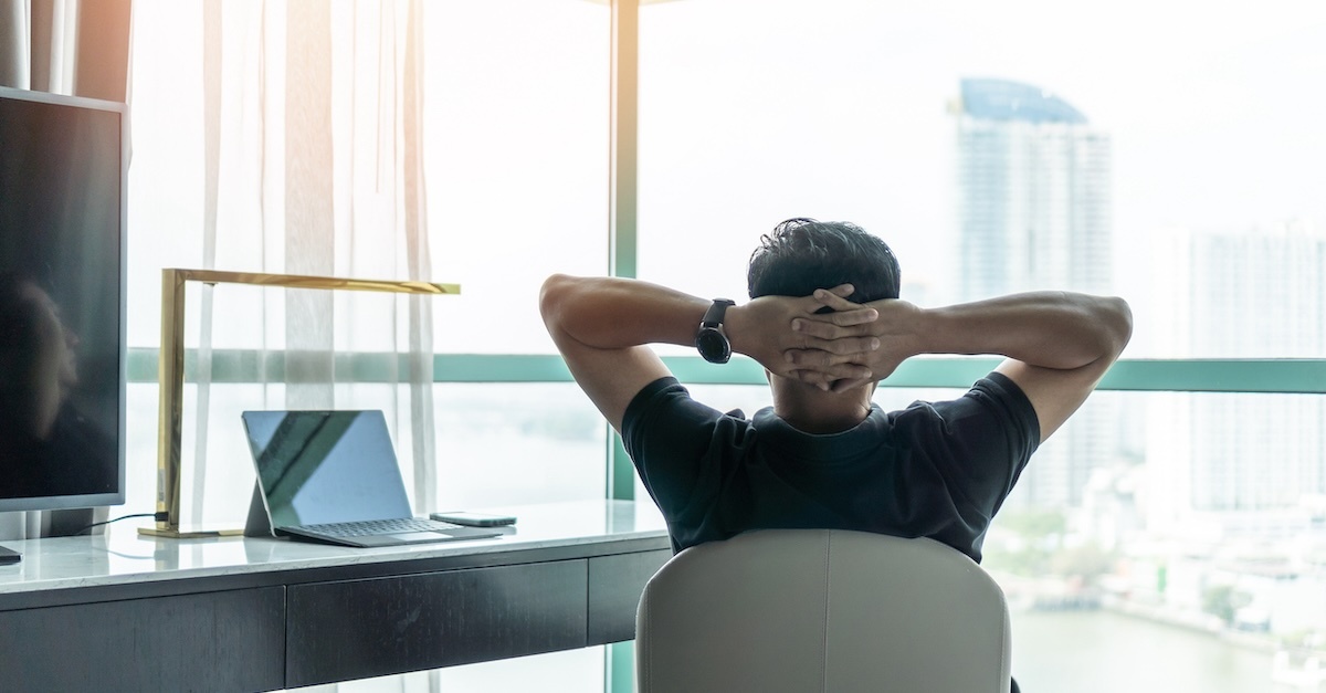 a man stretches his arms behind his head while looking out an office window and sitting at a desk with a laptop on it