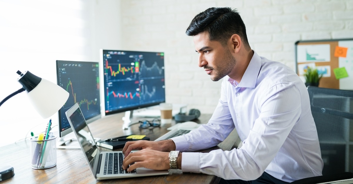 A man at his desk on a laptop with graphs on two extra screens