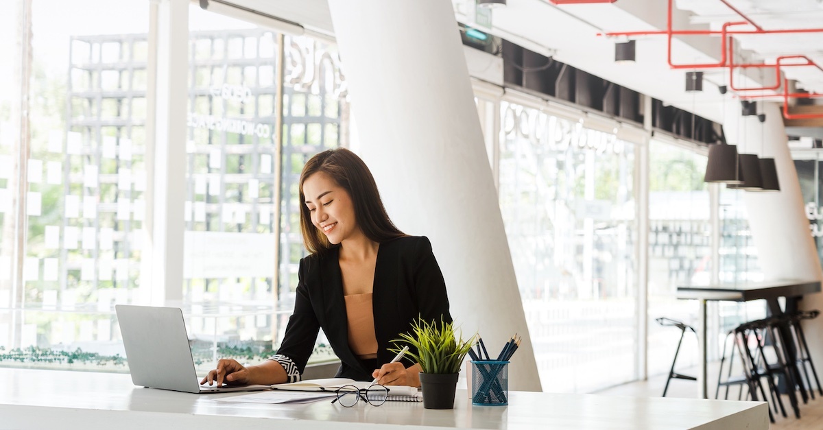 A woman in a professional setting smiles while working on her laptop in a modern, well-lit office space.