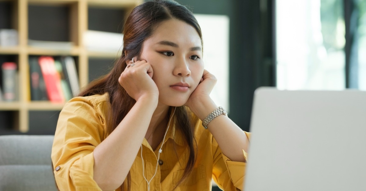 woman in a yellow blouse stressed over work in front of her laptop.