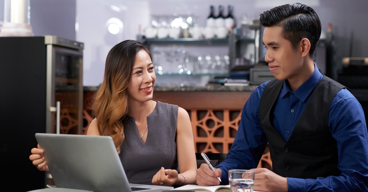 A man and woman work together in a restaurant with a laptop, a pen, and a notebook