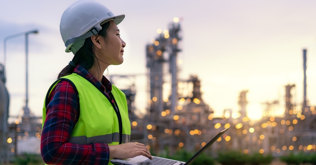 A woman in construction wearing a high-visibility vest and hard hat holds a laptop while looking over a construction site