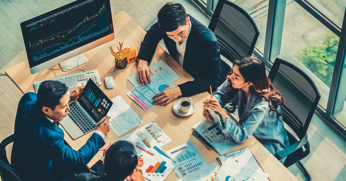 A group of professionals collaborates in a meeting room, reviewing documents and data charts on a table, with a large monitor displaying stock market information in the background