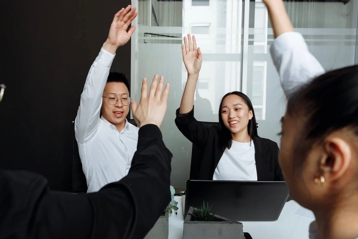 Dua orang karyawan sedang mengangkat tangan untuk meminta kesempatan berbicara pada sesi brainstorming di kantor.