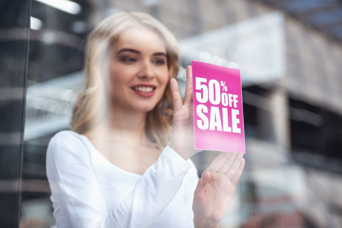 Seorang karyawan wanita sedang menempel contoh iklan penawaran diskon di kaca toko. (Sumber: Envato)