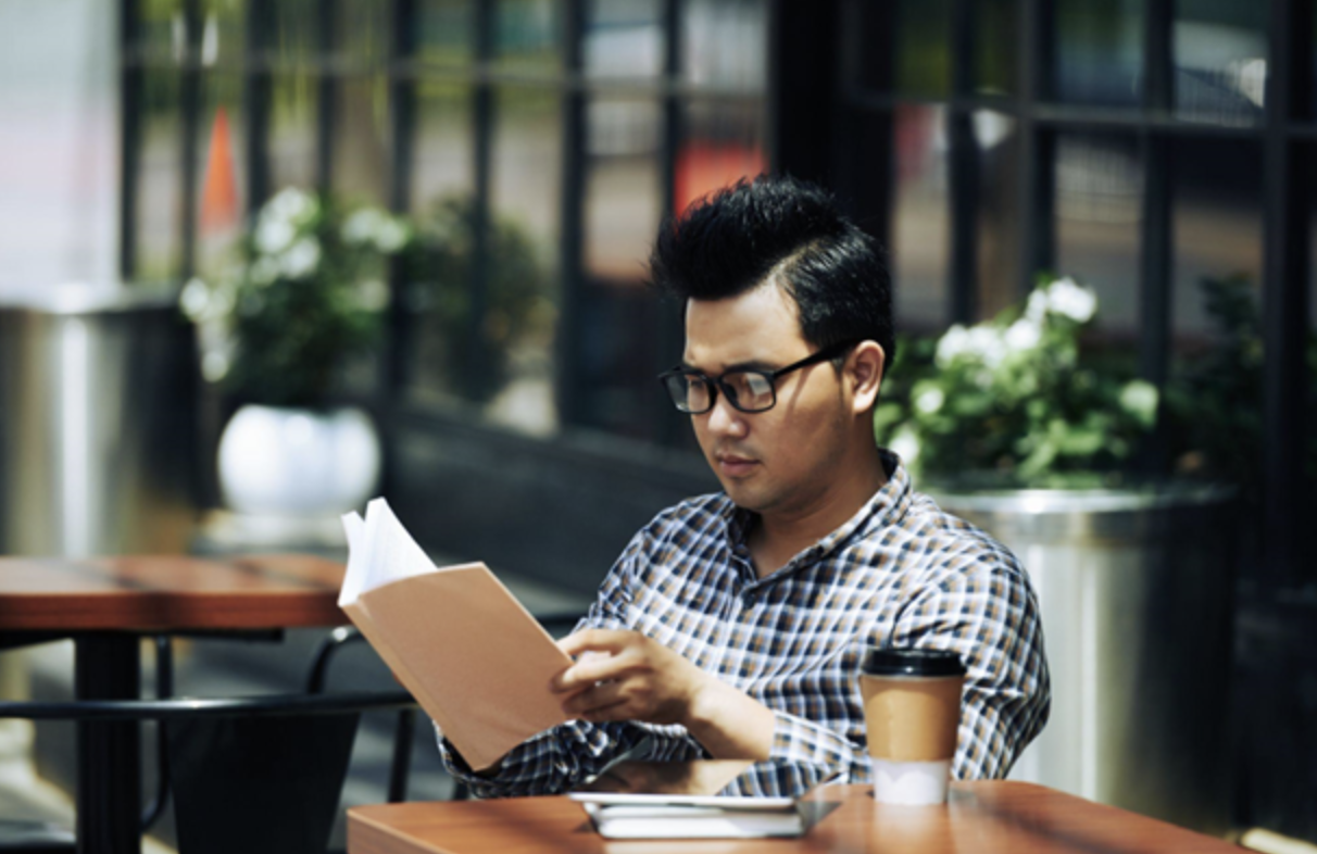 Young Asian man in glasses sitting at an outdoor cafe and reading book