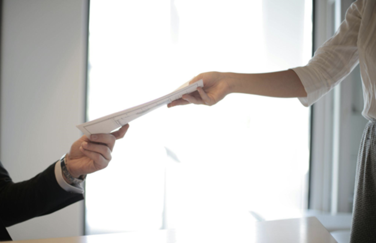 A woman in a white blouse hands a document to a man in a black suit