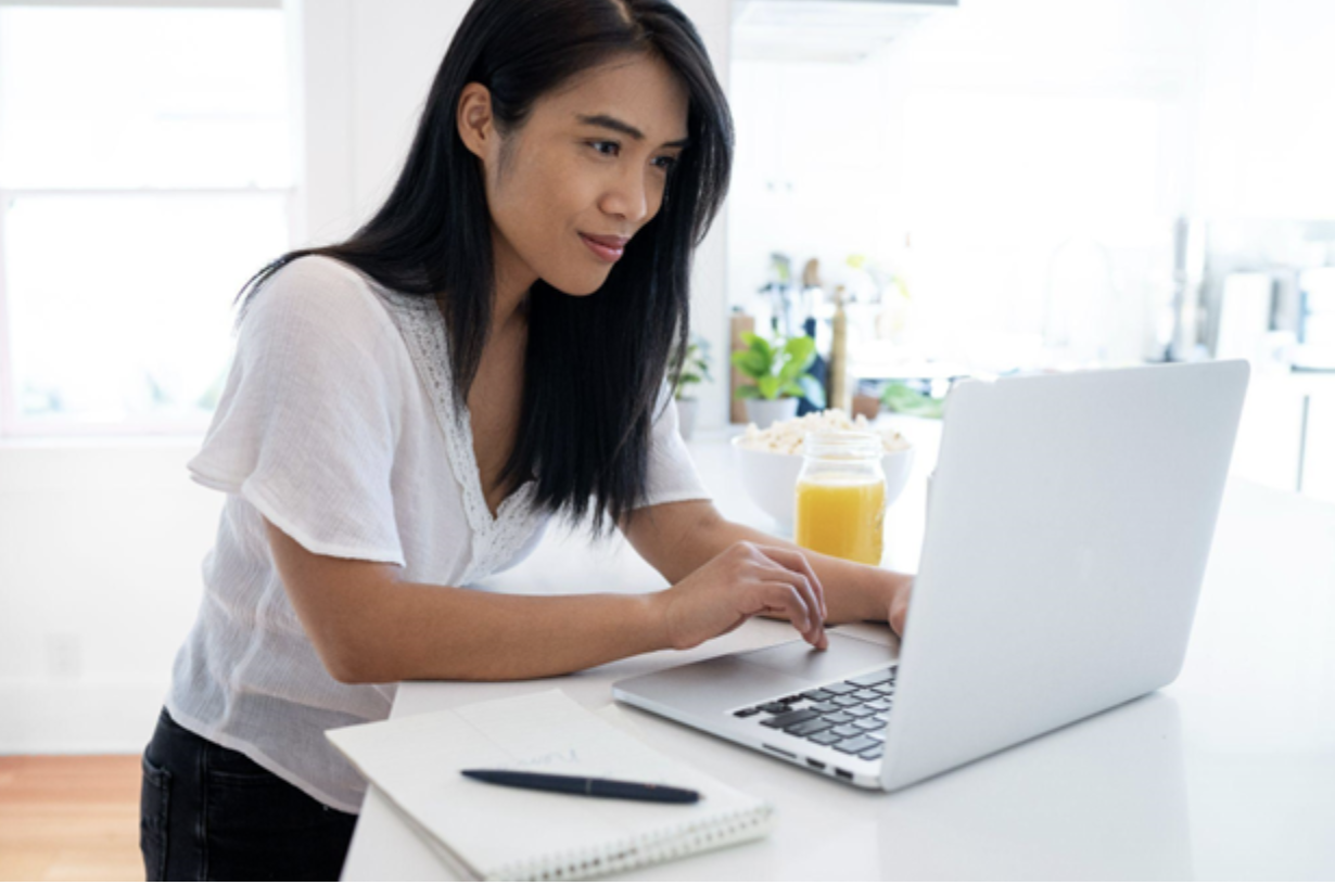 Young Asian woman typing on a laptop with a notebook and pen beside her