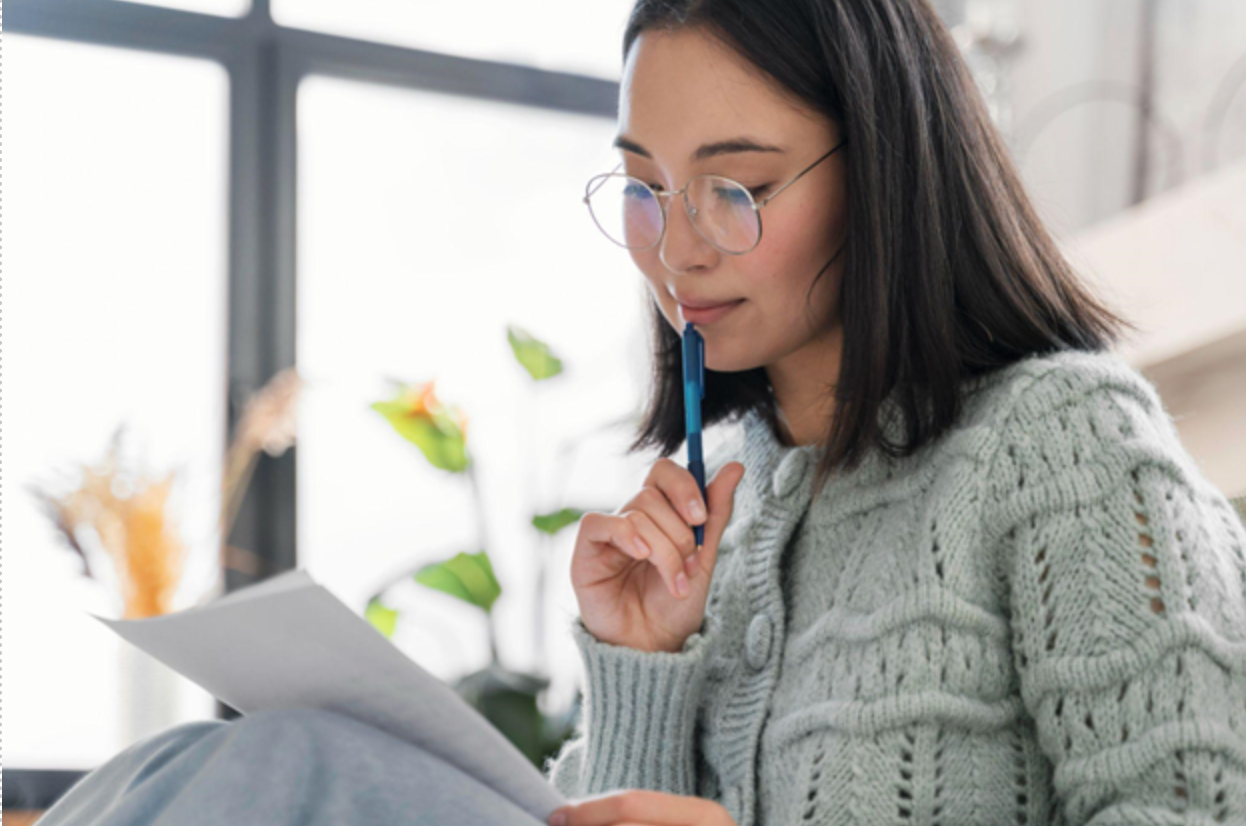 Young Asian woman in sweater looking thoughtfully at the letter, holding a pen to her chin