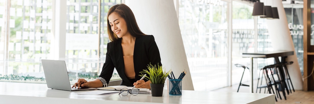 woman working at a table with a laptop, note pad, papers, glasses, pens, and a plant.