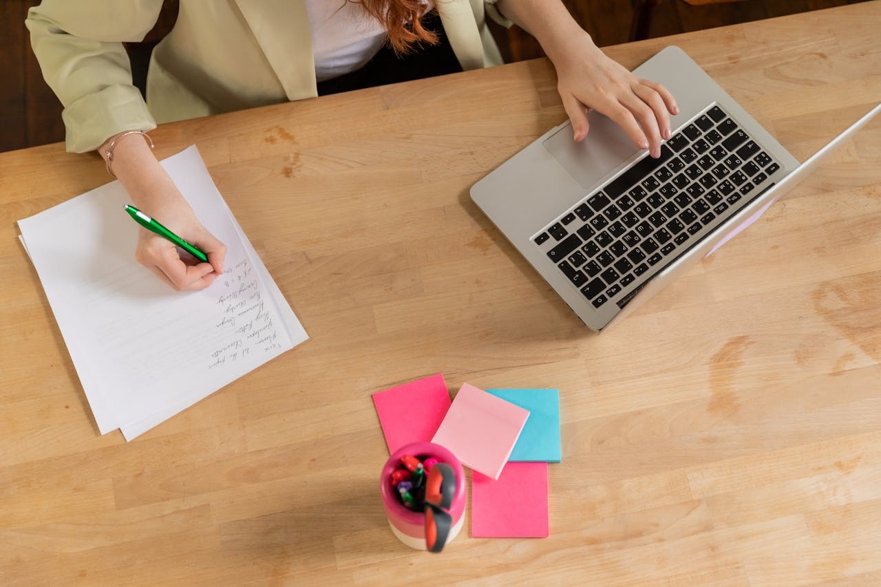 Mencatat poin-poin di atas kertas untuk membuat press release dengan tangan kanan sambil tangan kiri menyentuh keyboard laptop