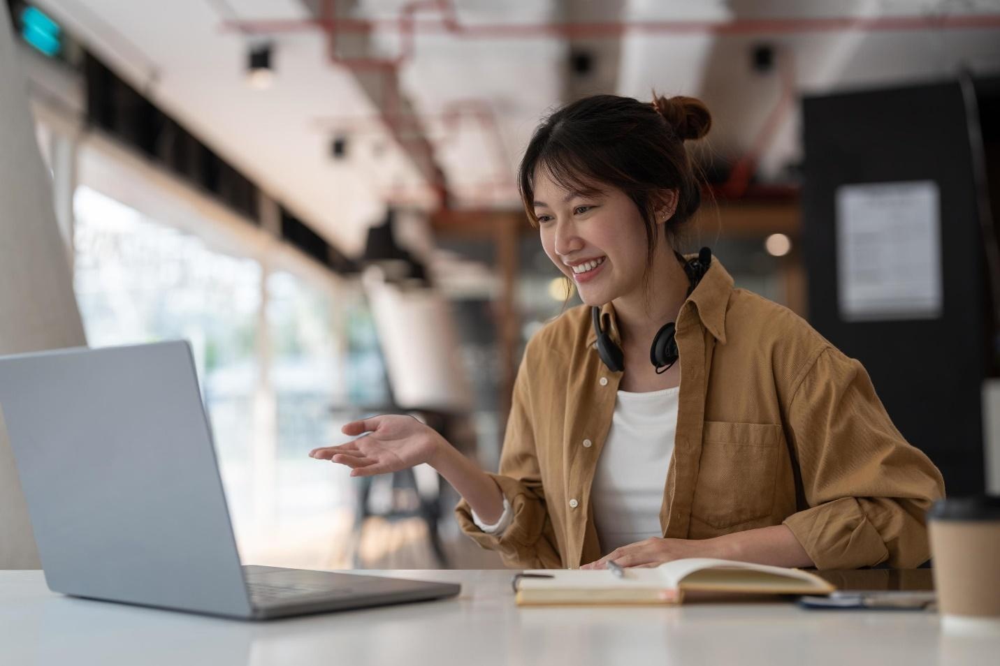 Young Asian woman smiling while using a laptop and taking notes in a modern workspace.