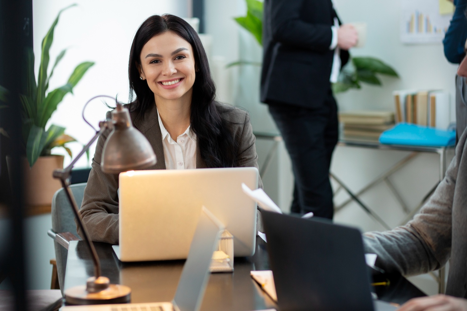 Wanita dengan outfit kerja blouse duduk tersenyum di depan laptop