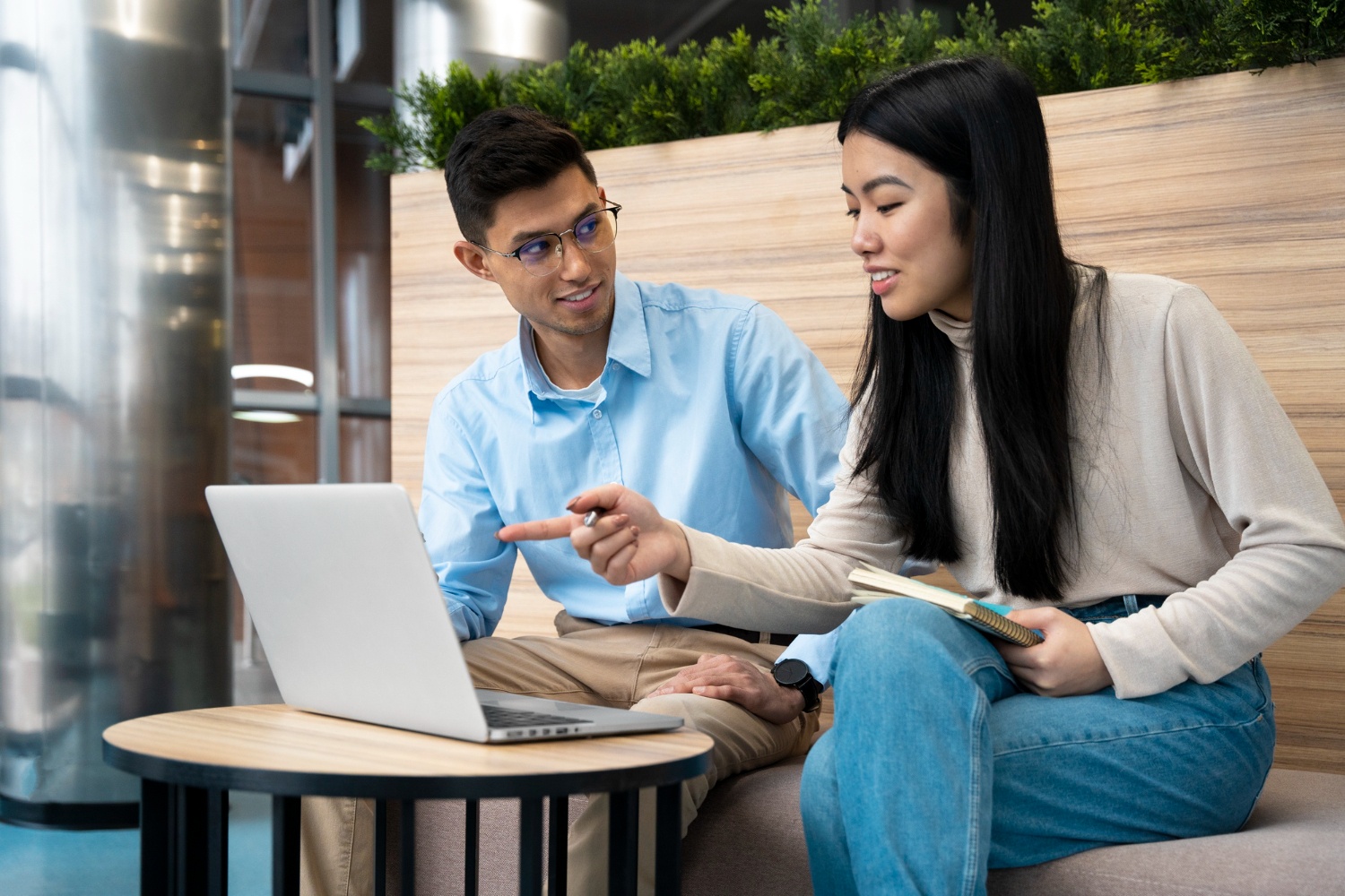 A young man and woman are sitting on a sofa, peering at a laptop. He's wearing glasses, and she's holding a notebook. They're both beaming. 