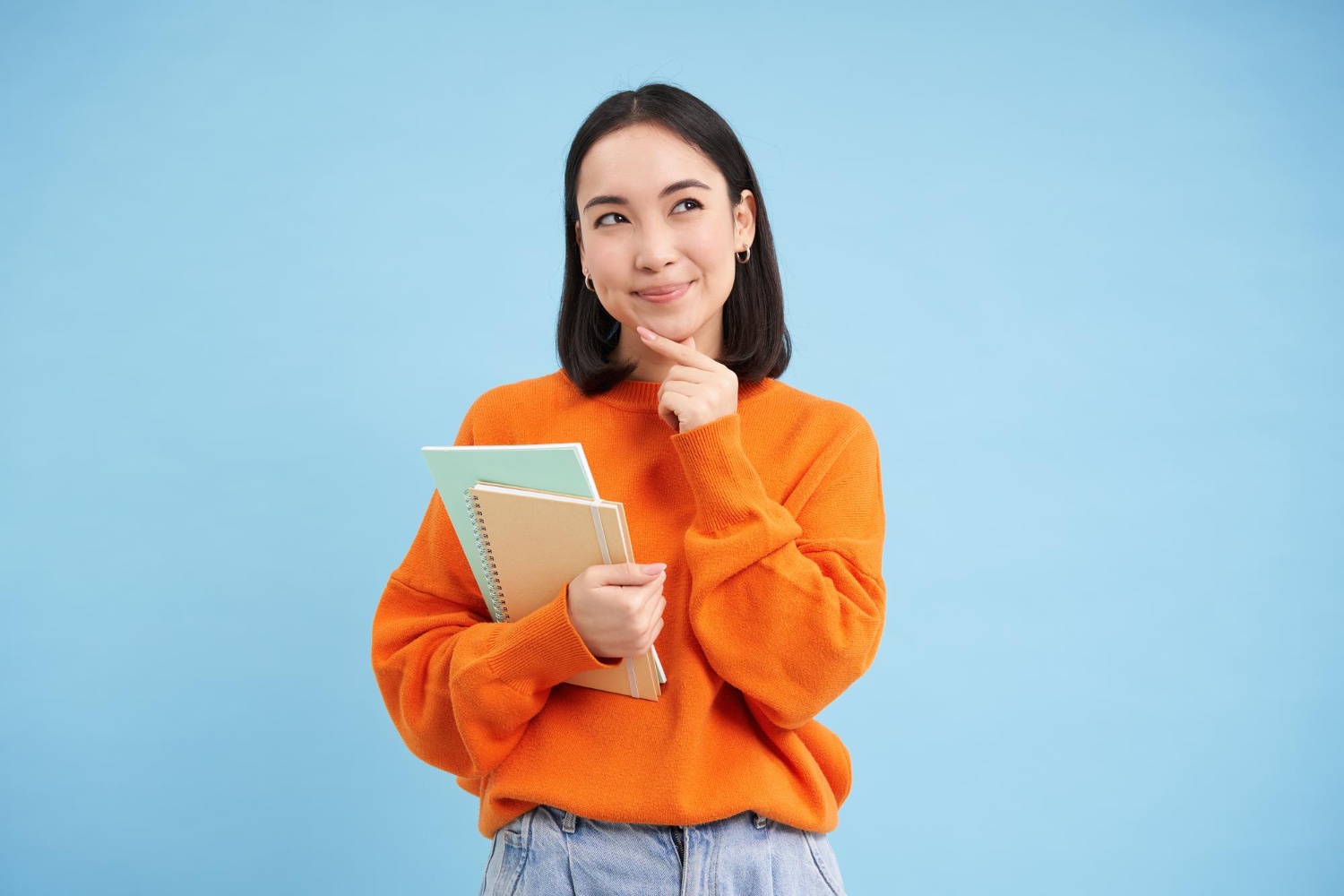 Thinking Asian girl holding two notebooks and looking upwards, against a blue background