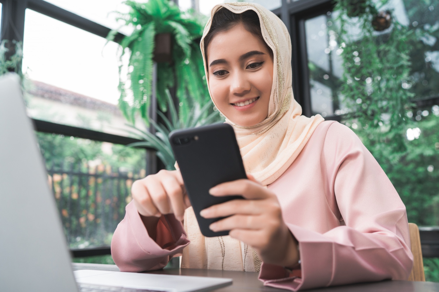 A young woman, her head wrapped in a colourful hijab, is sitting at a coffee shop table, engrossed in her phone. 