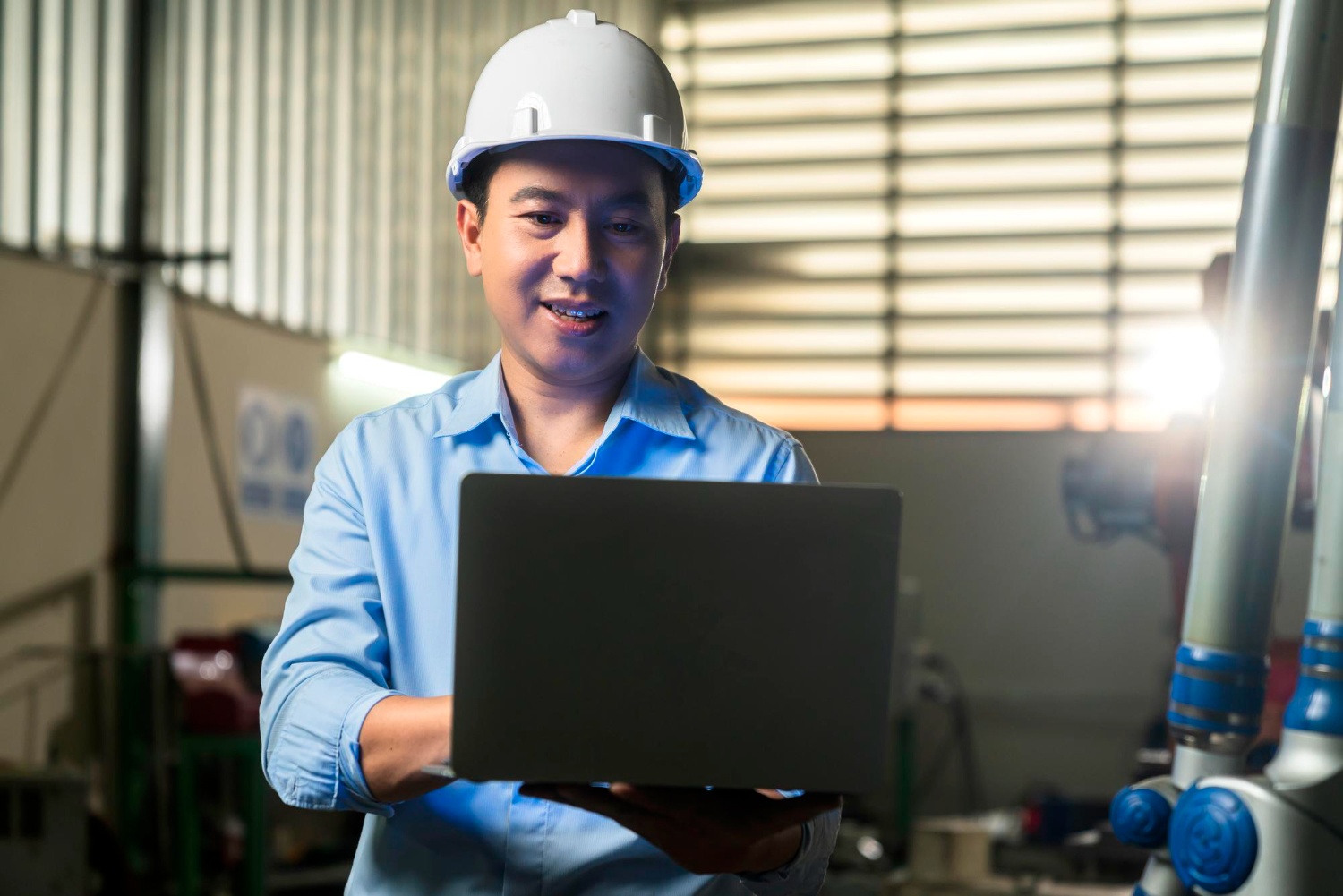 Pekerja perusahaan manufaktur berhelem putih sedang berdiri sambil memegang laptop terbuka