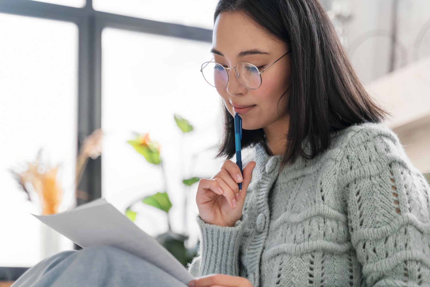 Young Asian woman in sweater looking thoughtfully at letter, holding a pen to her chin