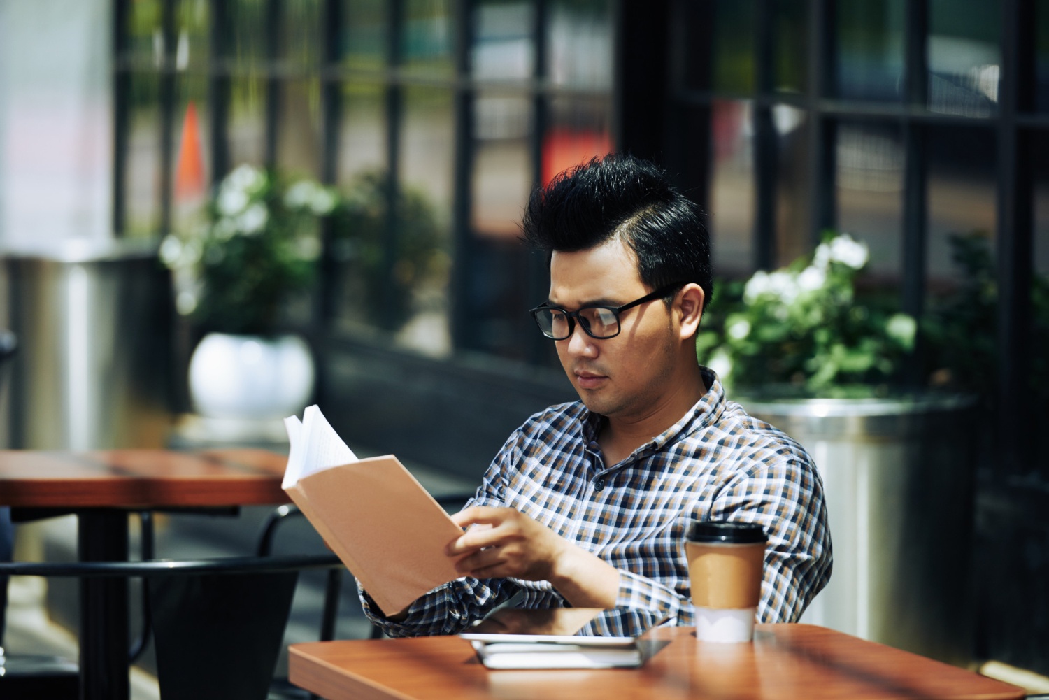 Young Asian man in glasses sitting at an outdoor cafe and reading book