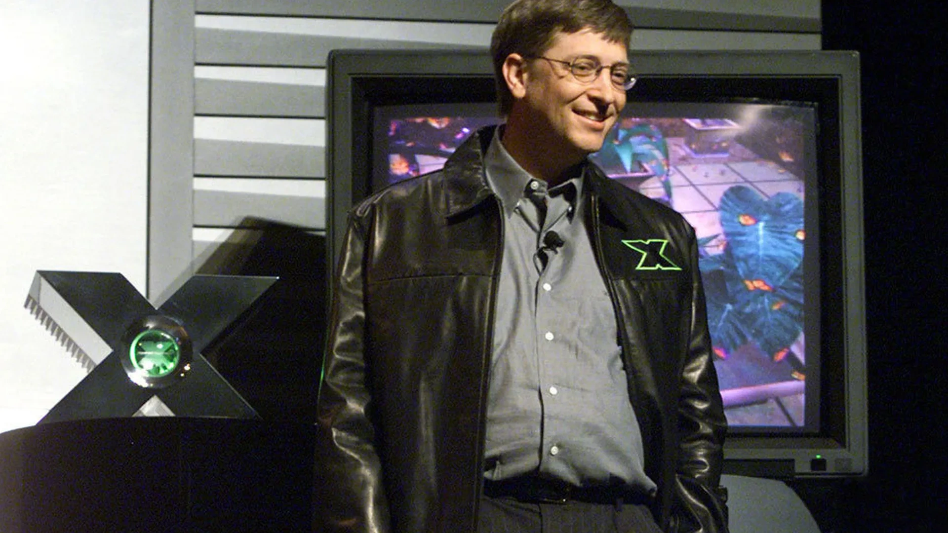 Protótipo do Xbox, feito de alumínio à esquerda, Bill Gates sorrindo à direita, e atrás dele uma televisão com uma imagem de uma demonstração do poder do console