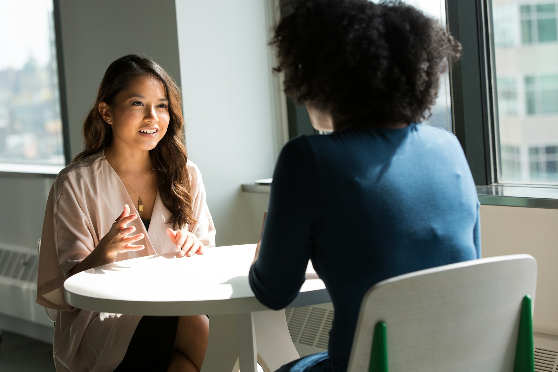 Two women sit across from each other at a table in a bright office, engaged in a discussion, possibly about providing a personal reference for a job application