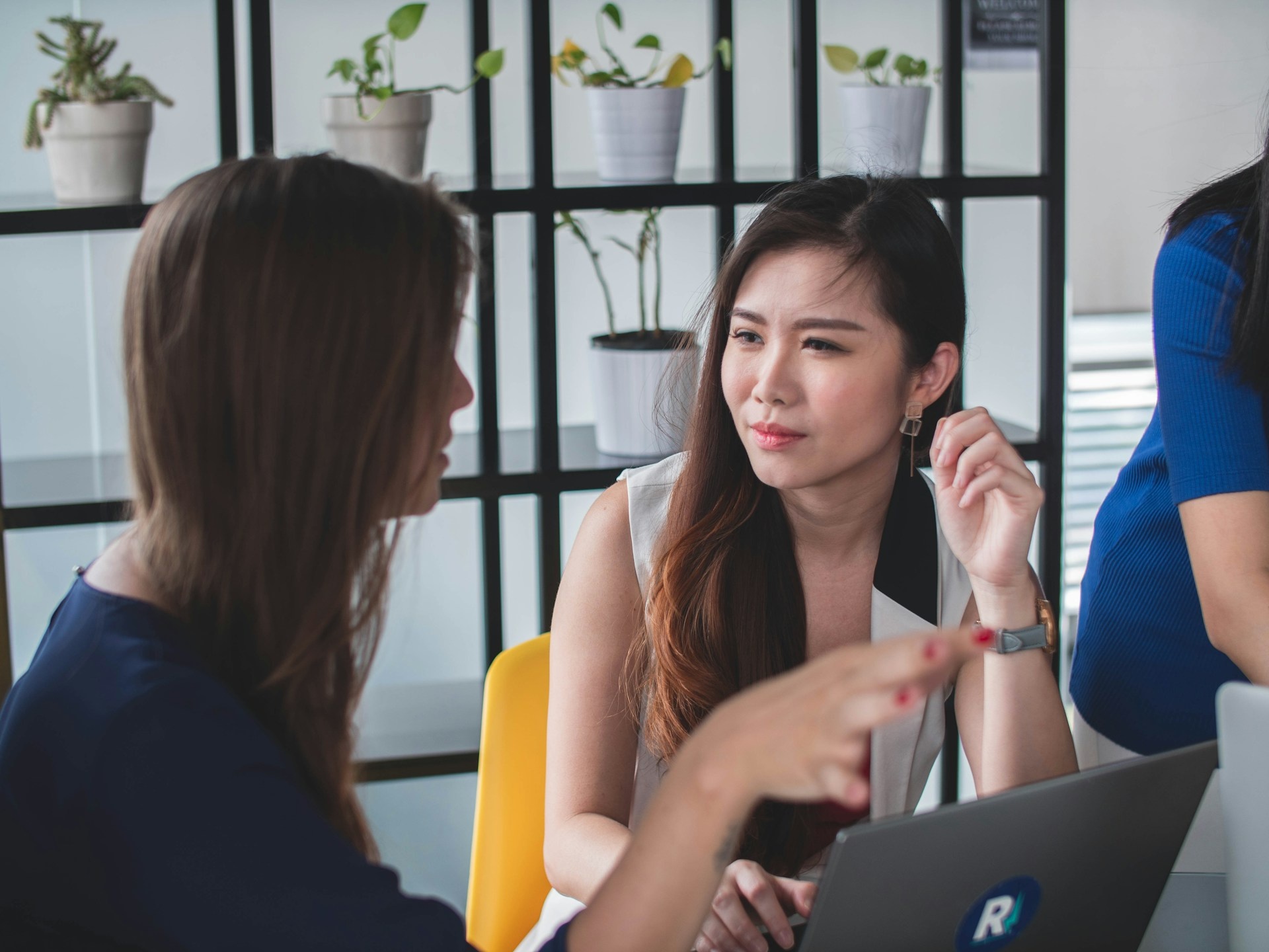 Two executives engage in a serious discussion at a desk, possibly about providing a personal reference, with a laptop and potted plants in the background