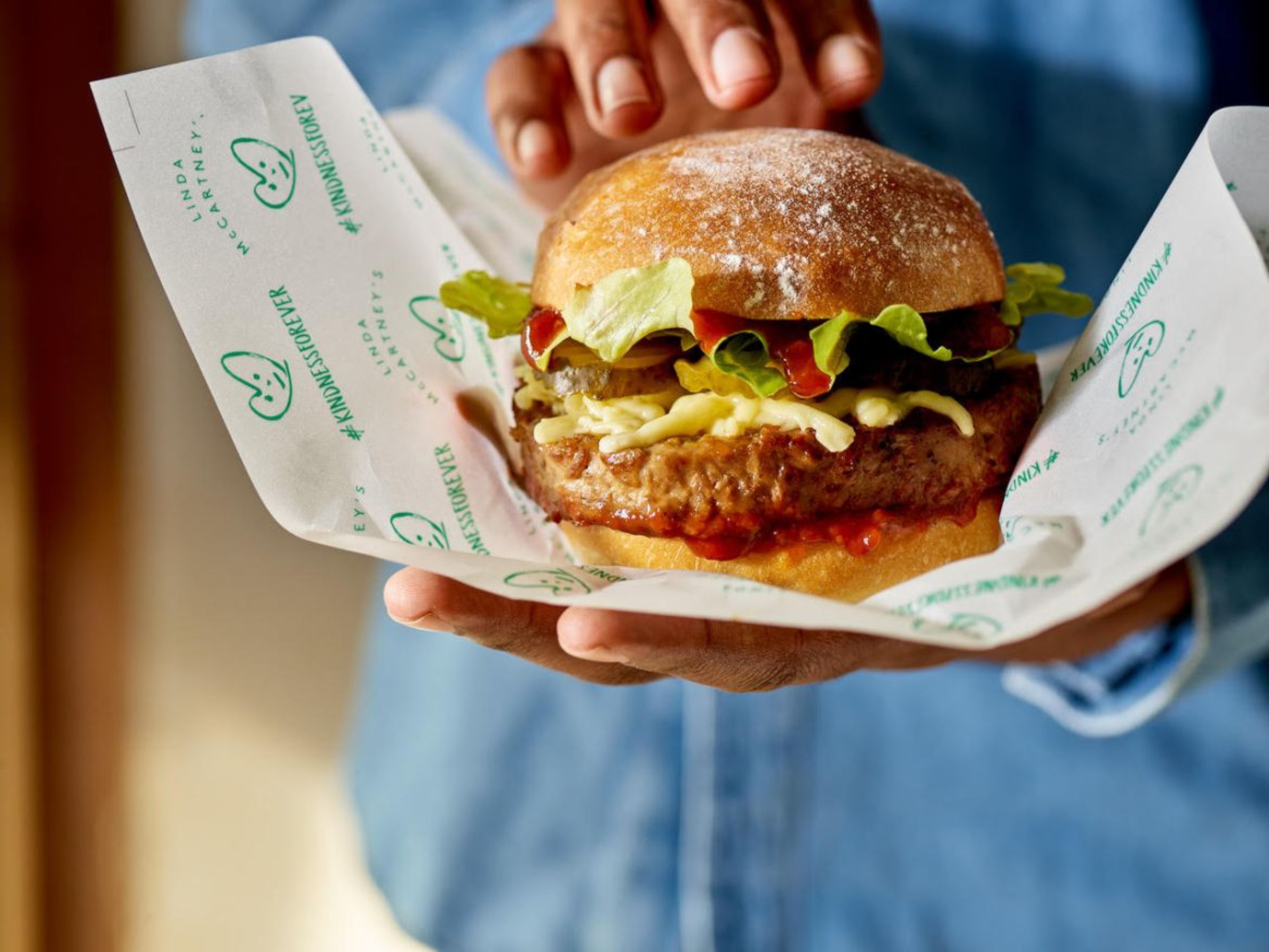A hand holding a burger in a bun with cheese and lettuce, wrapped in paper