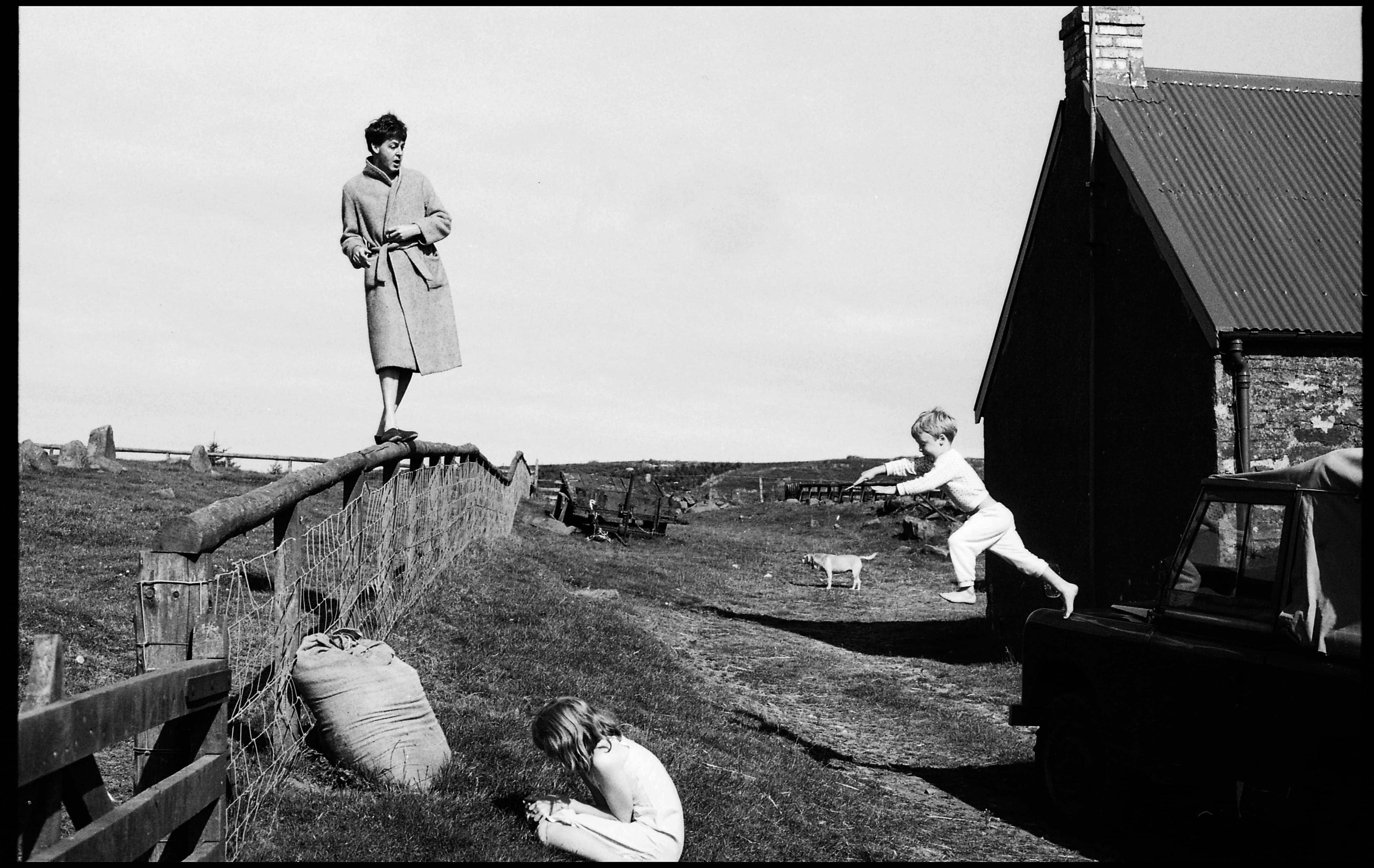 Black and white photo of Paul standing on a fence in a farmyard, while James jumps in the air