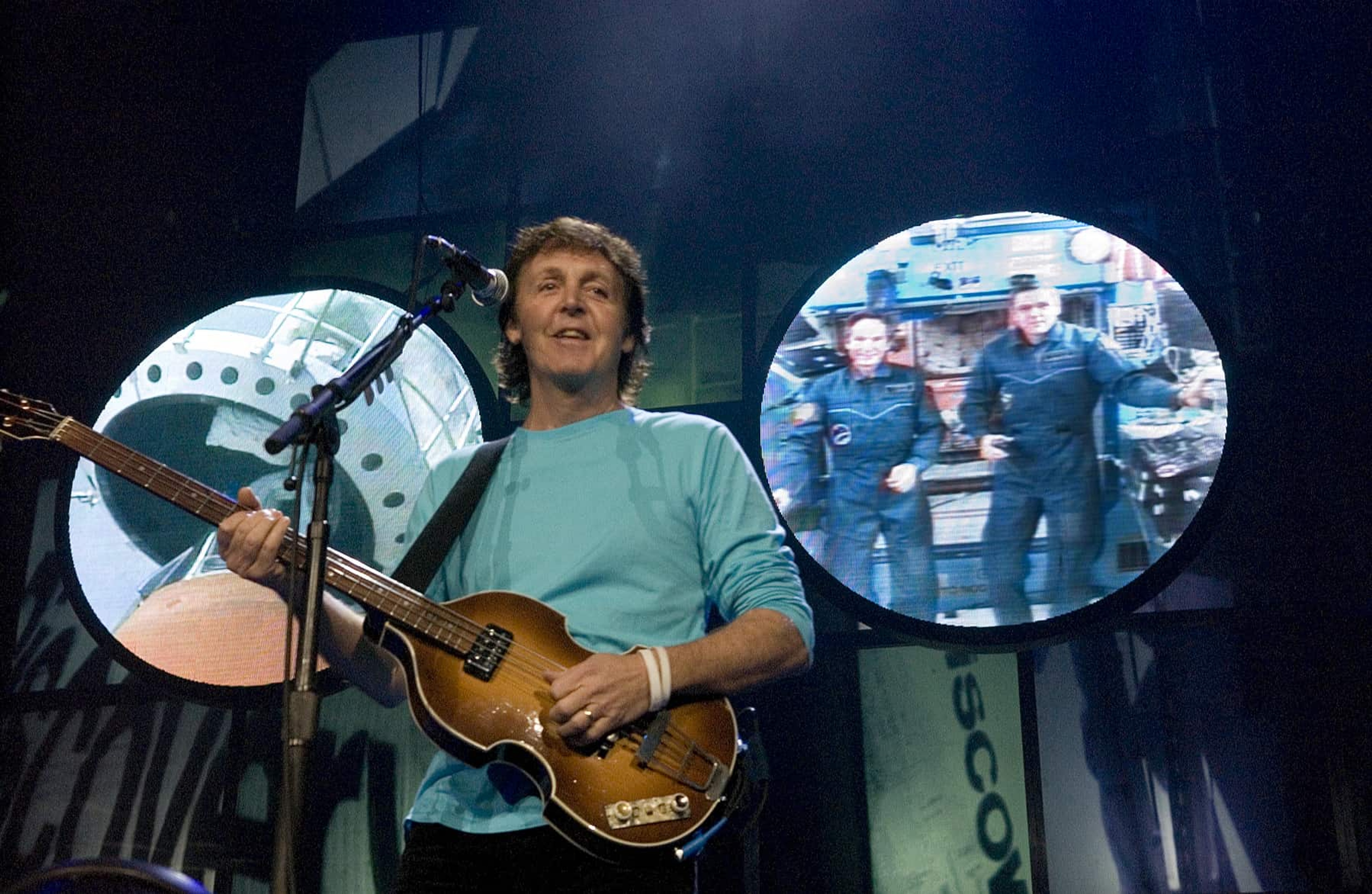 Paul on stage holding a guitar, with astronauts on a screen behind him